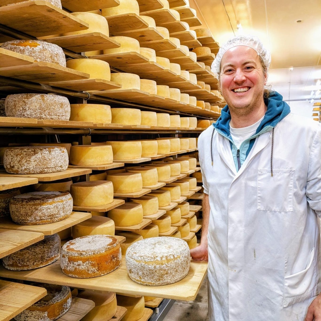 Porträt von Thilo Metzger-Petersen im Käselager des Backensholzer Hofs. Er steht zwischen gestapelten Käselaiben, lacht offen in die Kamera.