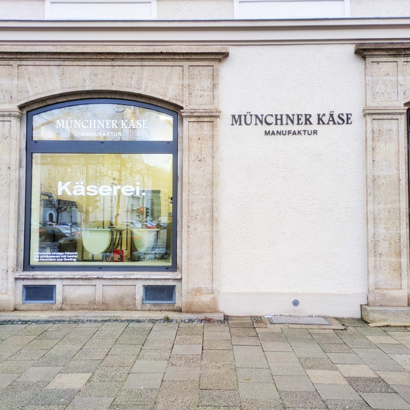 Außenansicht der Münchener Käsemanufaktur in München mit großer Fensterfront. Durch die Glasfassade ist die gläserne Käserei sichtbar
