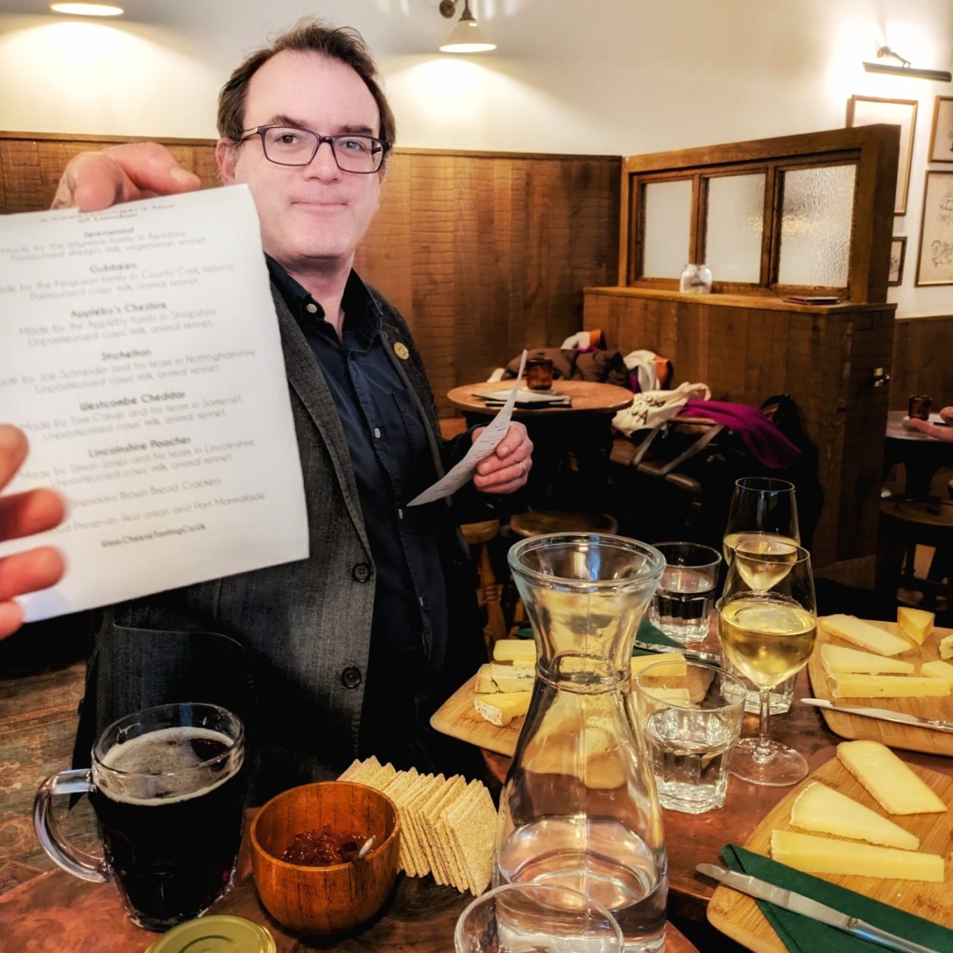 Ned Palmer sitzt in einem Londoner Pub vor einer vorbereiteten Käseauswahl – aufgeschnittene Stücke auf Holzbrett, Pintglas daneben.
