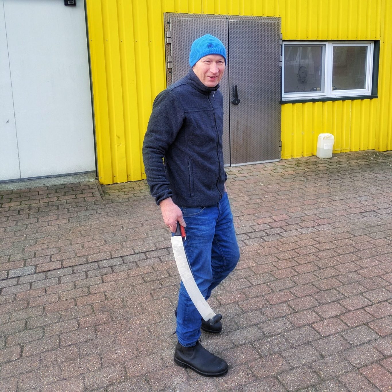 Hauke Koll vor der gelben Trapezblechfassade der Ostenfelder Meierei Hauke Koll mit blauer Mütze geht vor einer gelben Trapezblechfassade und hält ein Käsemesser in der Hand.