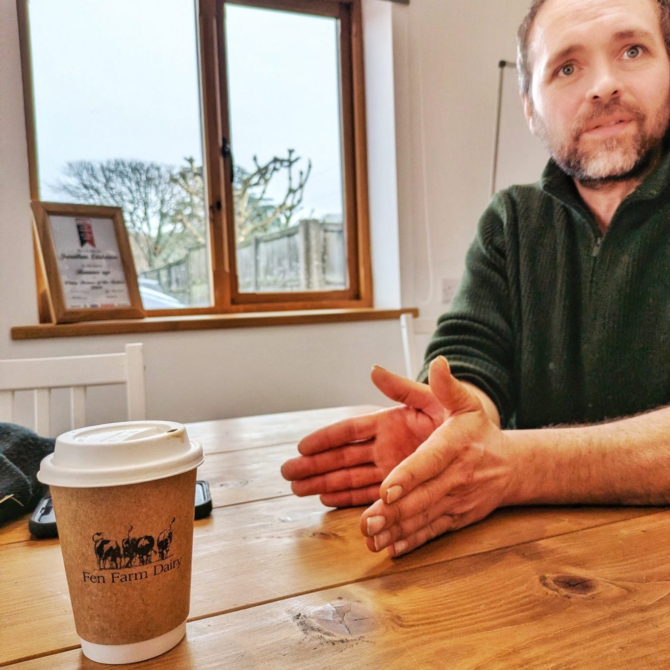 Jonny Crickmore im Gespräch, sitzend an einem Holztisch im Meetingraum der Fen Farm Dairy in Suffolk, England.